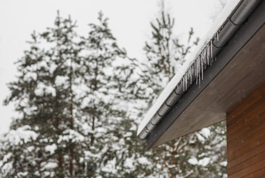 The side of a home in winter with a propane tank nearby, gutters lined with snow and ice hanging down.
